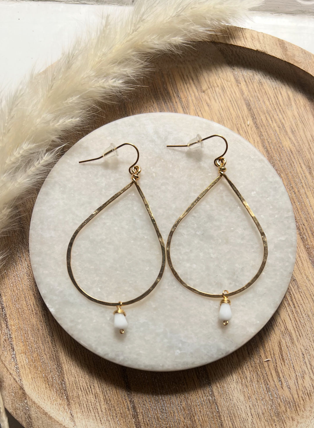 Gold hammered teardrop earrings with small white bead drop, displayed on a round marble tray with wooden background.