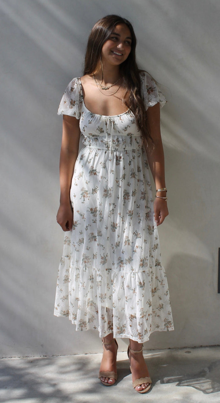 Woman in a white floral dress standing against a light gray wall.