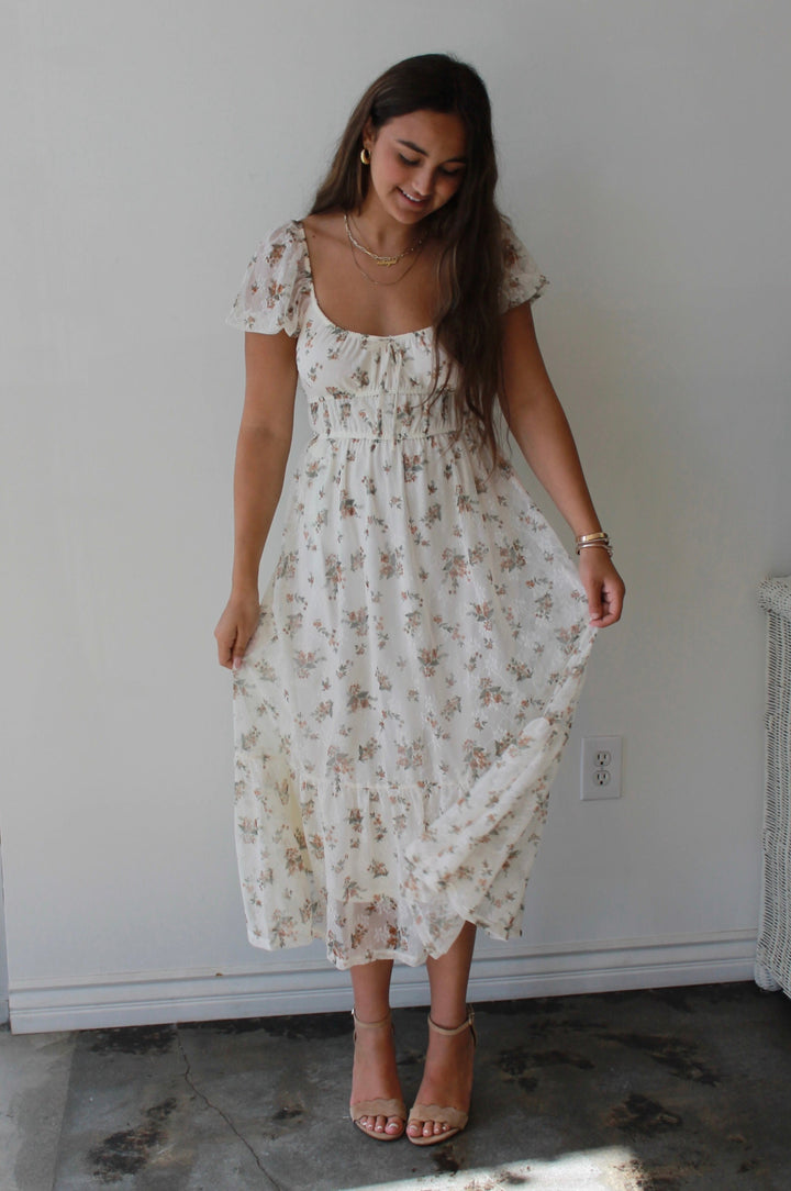 Woman wearing a floral dress standing in a room with a white wall and a window.