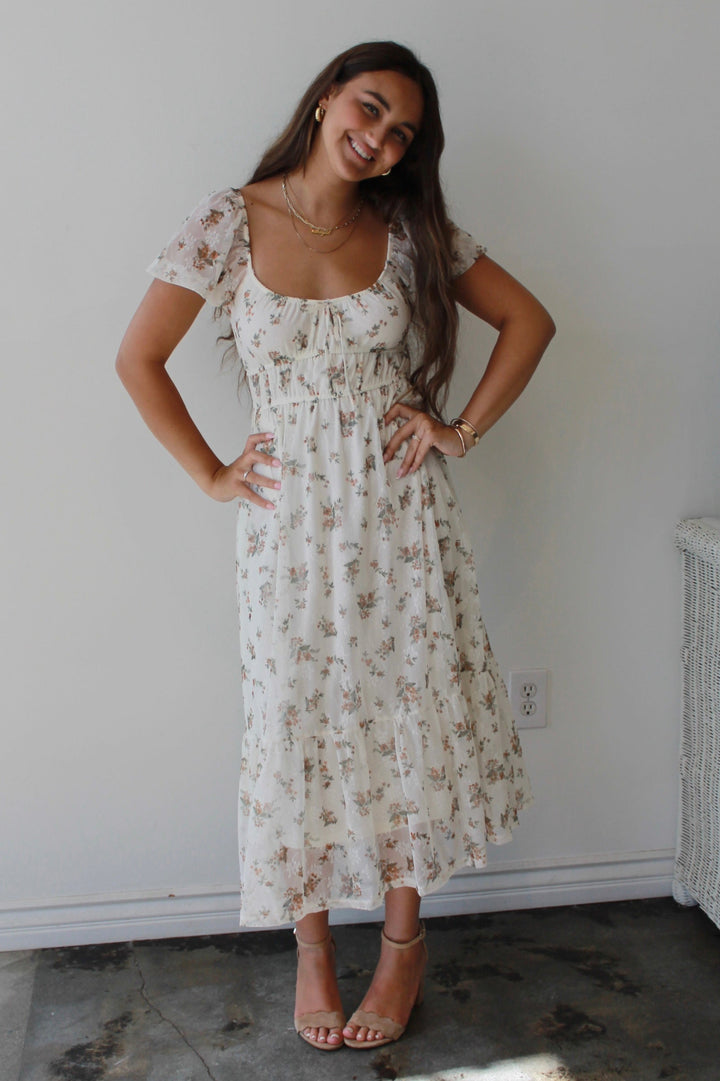 Woman wearing a floral dress standing in a room with a white wall and a window.