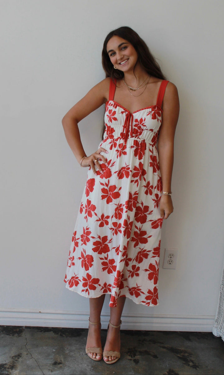 Woman wearing a white dress with red floral patterns standing against a white wall.