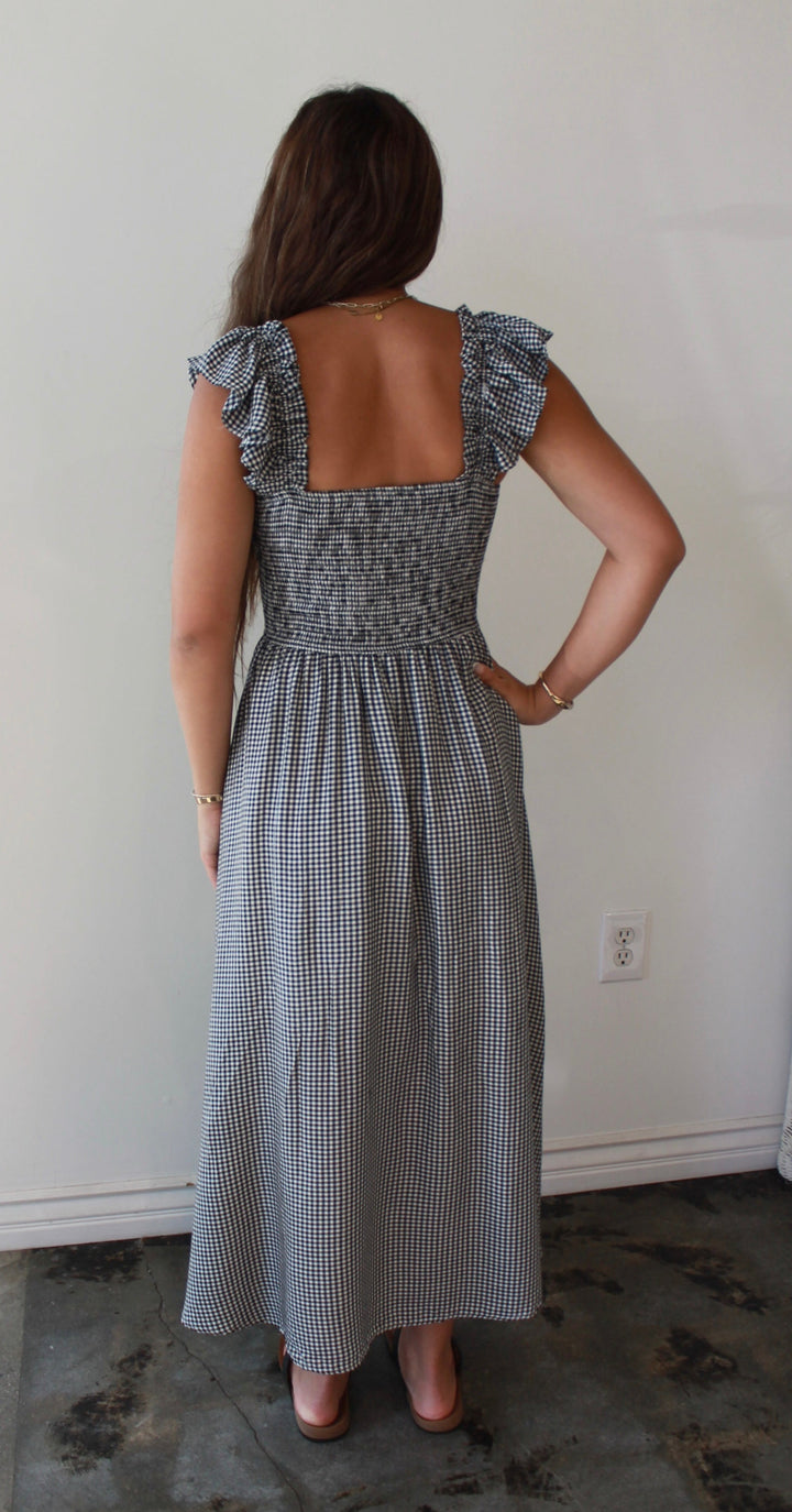 Woman wearing a checkered dress standing in a room with a white wall and concrete floor.