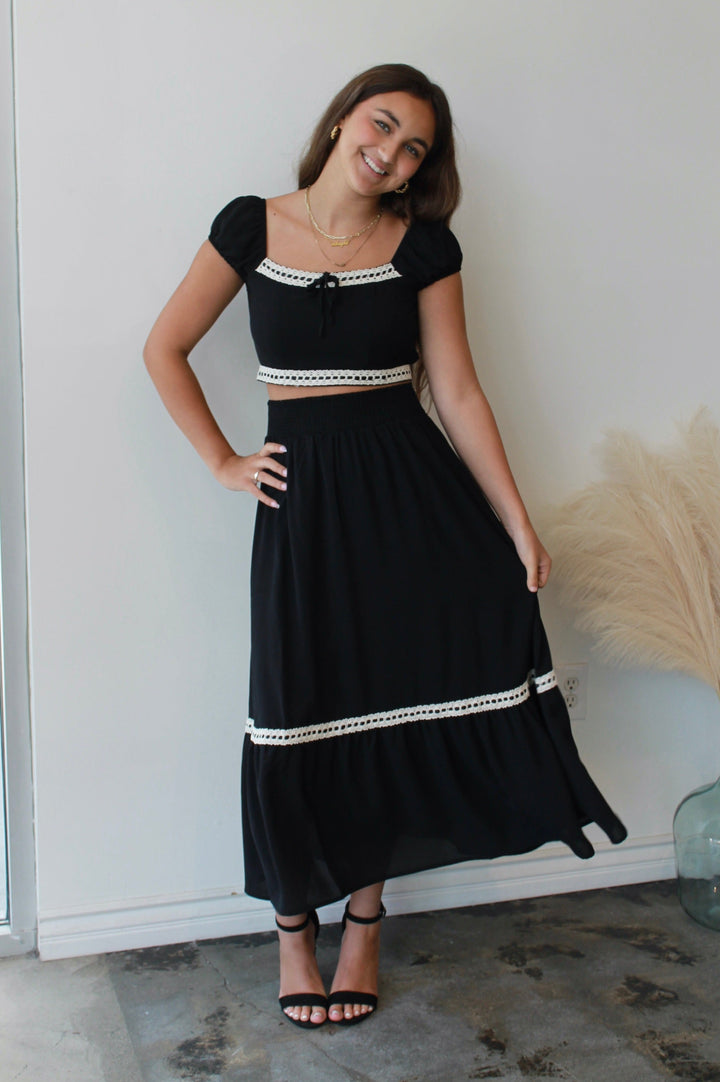 Woman wearing a black top and skirt with white trim standing indoors.