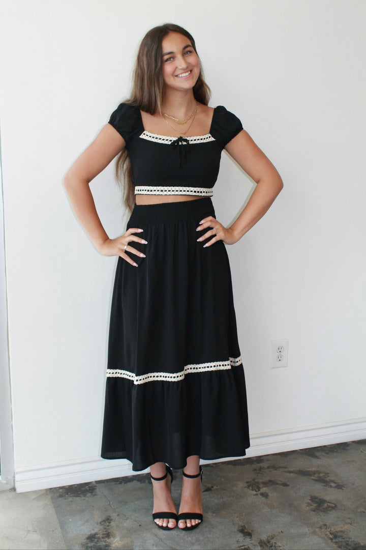 Woman wearing a black skirt set with white trim against a white wall.