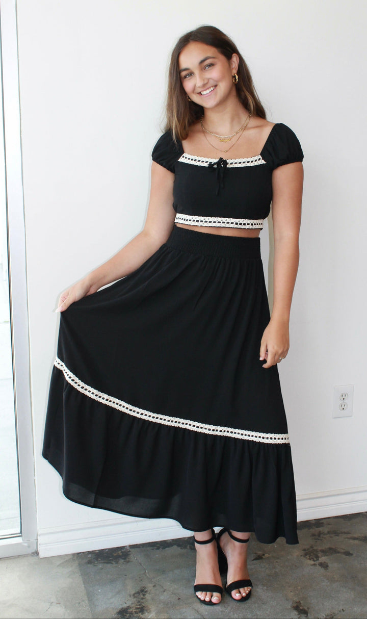 Woman wearing a black skirt set with white trim against a white wall.