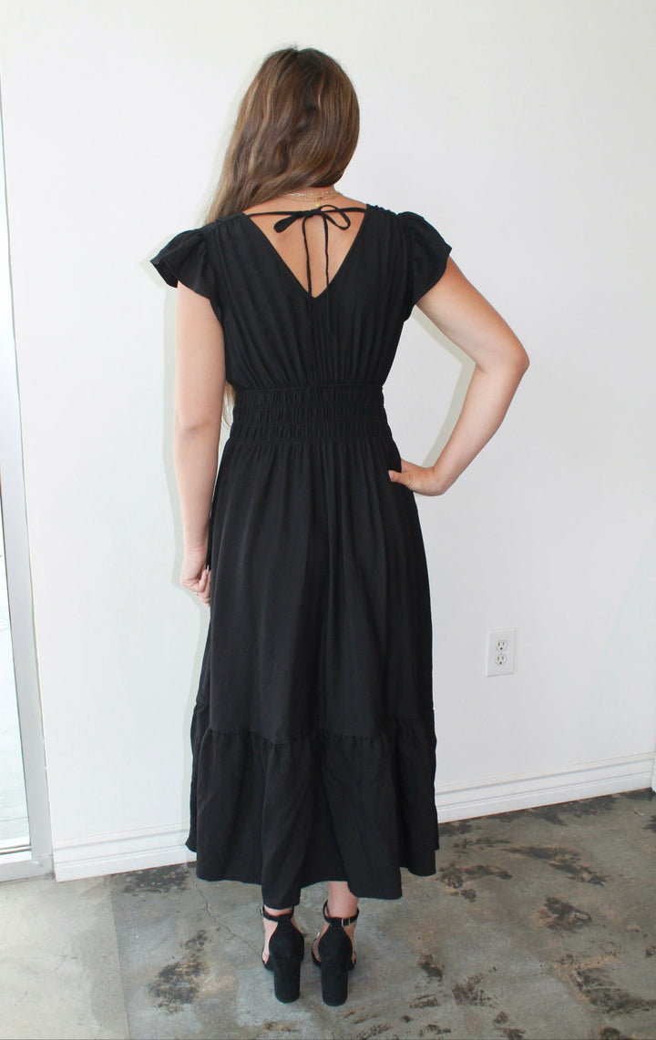 Woman wearing a black dress with a back bow detail in a minimal indoor setting.
