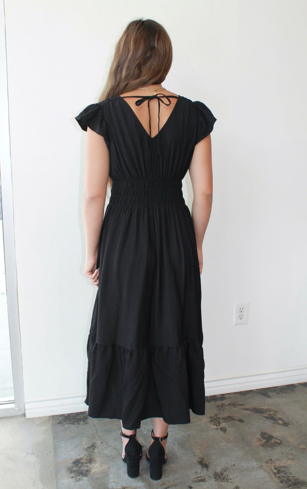 Woman wearing a black dress standing in a room with a white wall and concrete floor.
