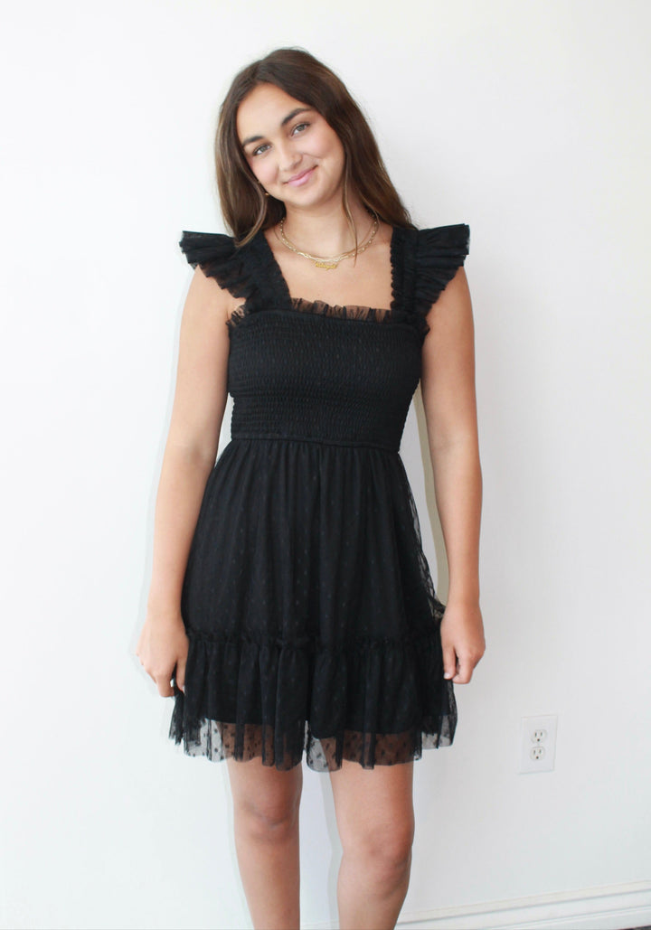 Woman wearing a black dress with ruffled shoulders against a white wall.