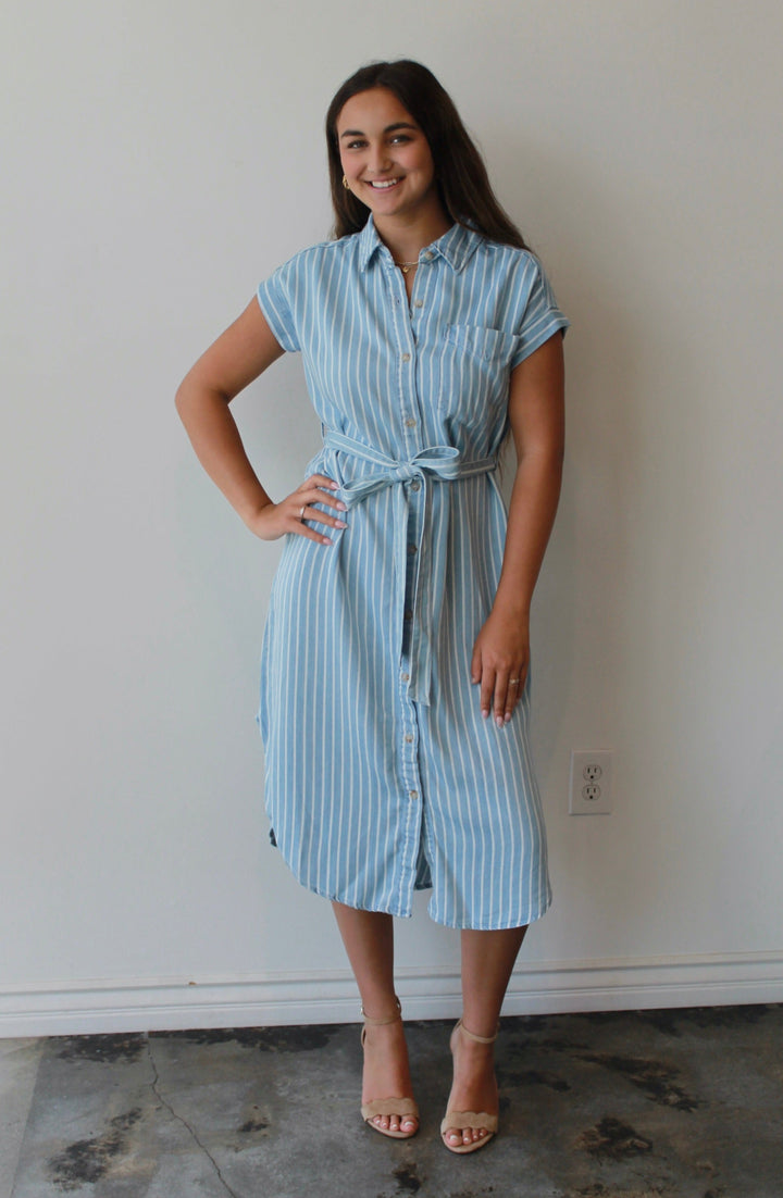 Woman wearing a light blue striped dress standing against a white wall.