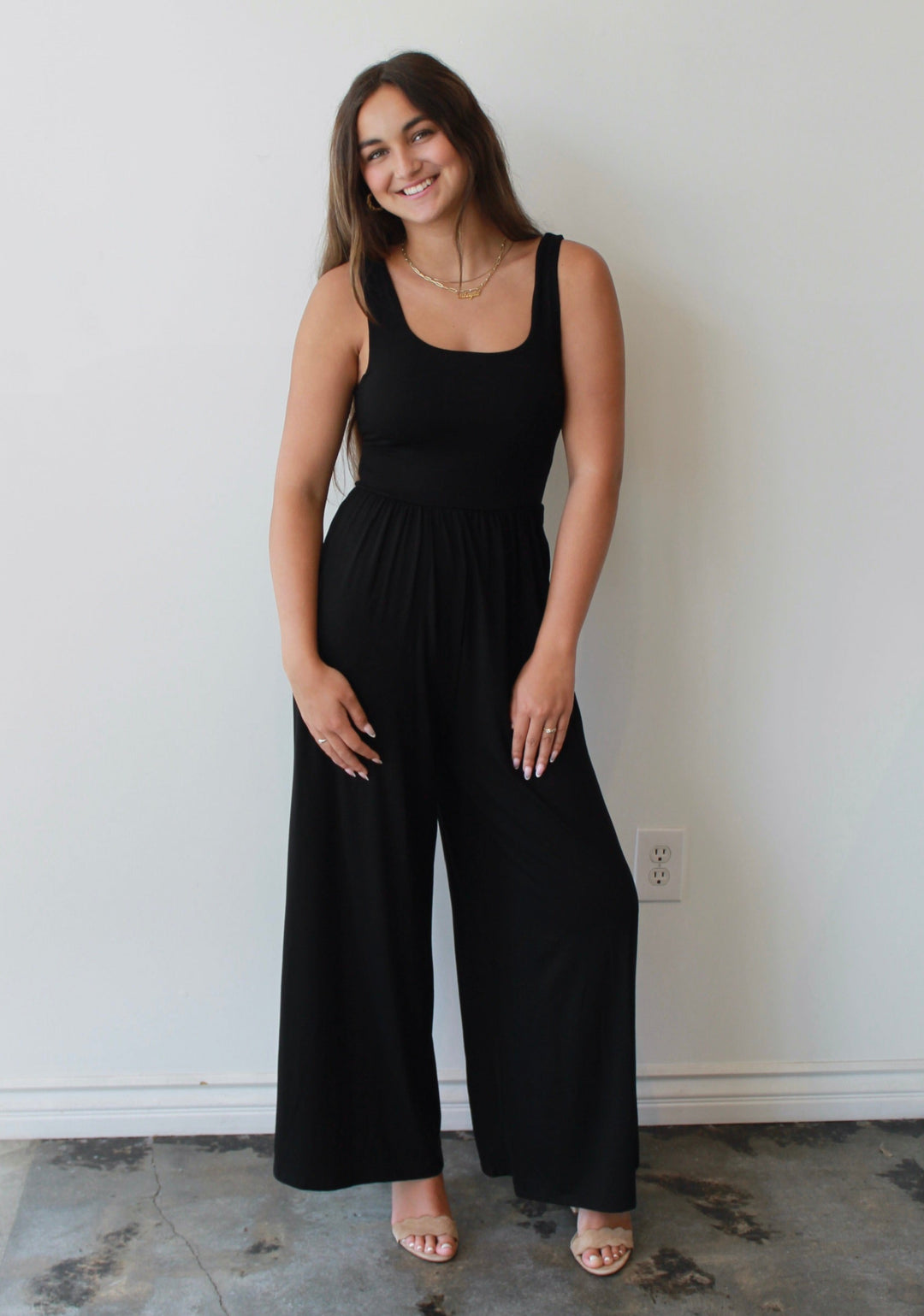 Woman wearing a black jumpsuit standing against a white wall.