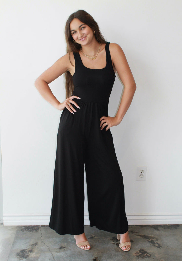 Woman wearing a black jumpsuit standing against a white wall.