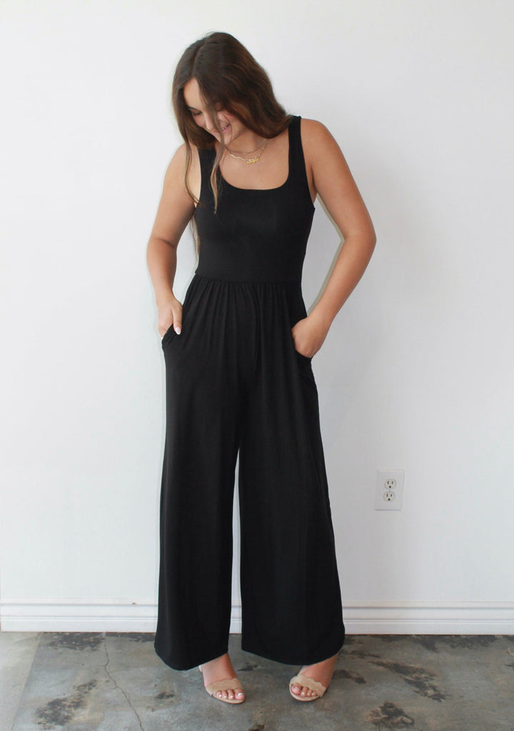 Woman wearing a black jumpsuit standing against a white wall.