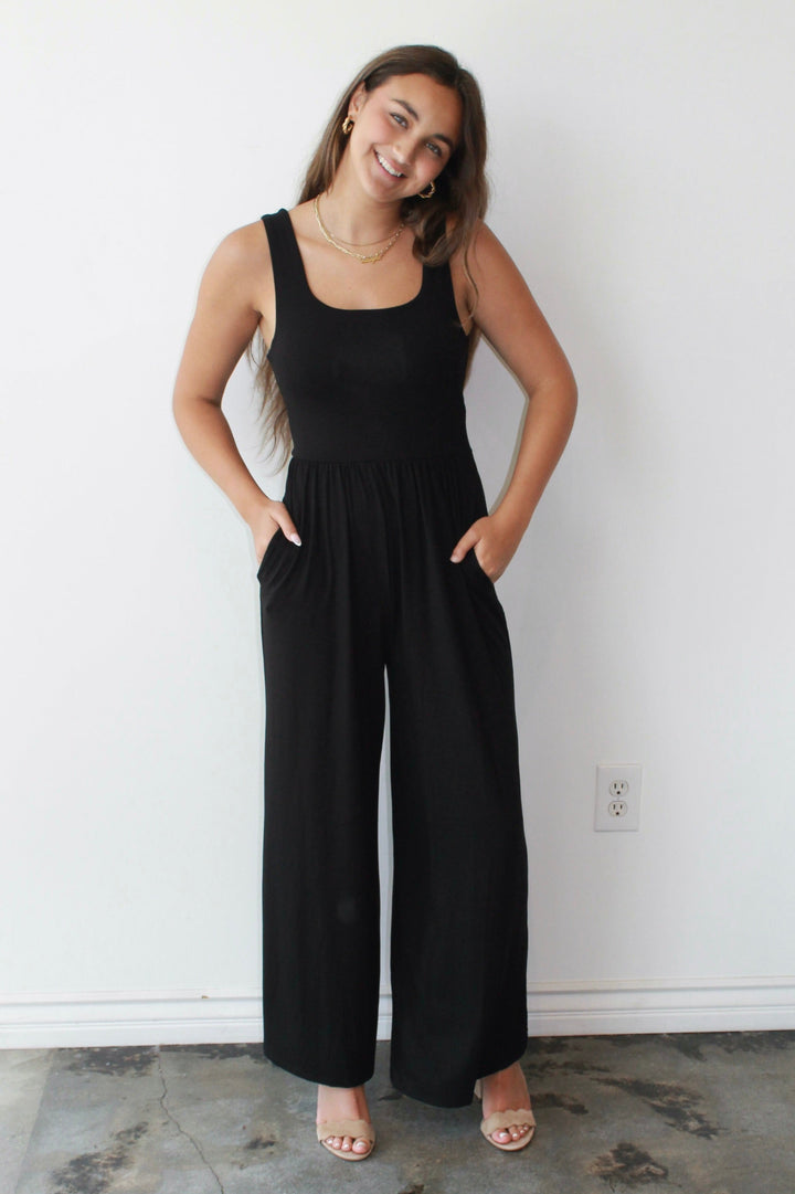 Woman wearing a black jumpsuit standing against a white wall.