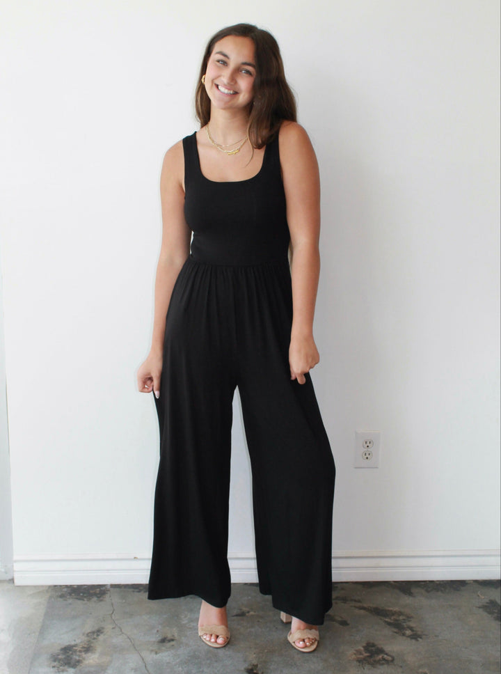 Woman wearing a black jumpsuit standing against a white wall.