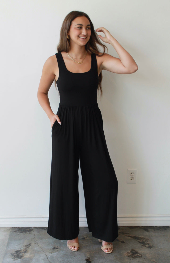 Woman wearing a black jumpsuit standing against a white wall.