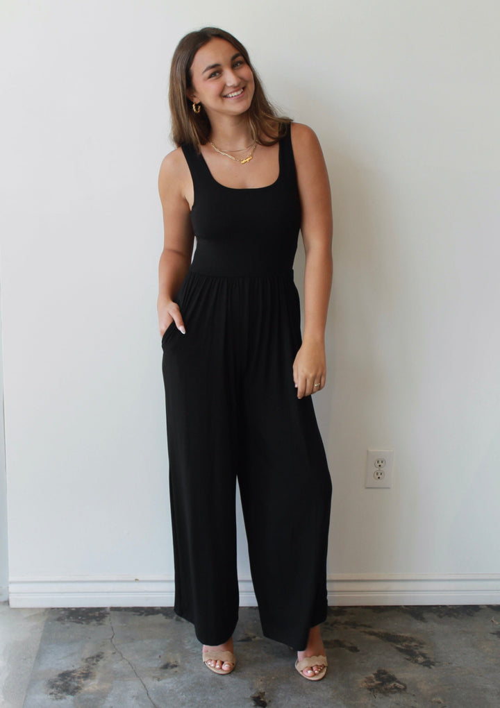 Woman wearing a black sleeveless jumpsuit standing against a white wall.