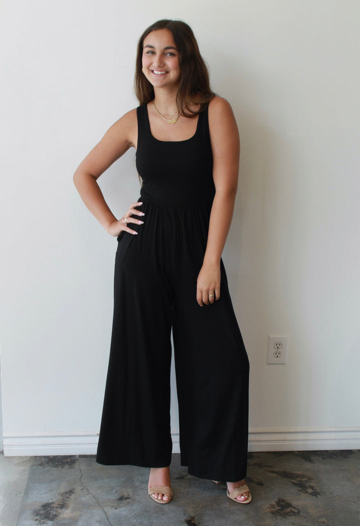 Woman wearing a black jumpsuit standing against a white wall.
