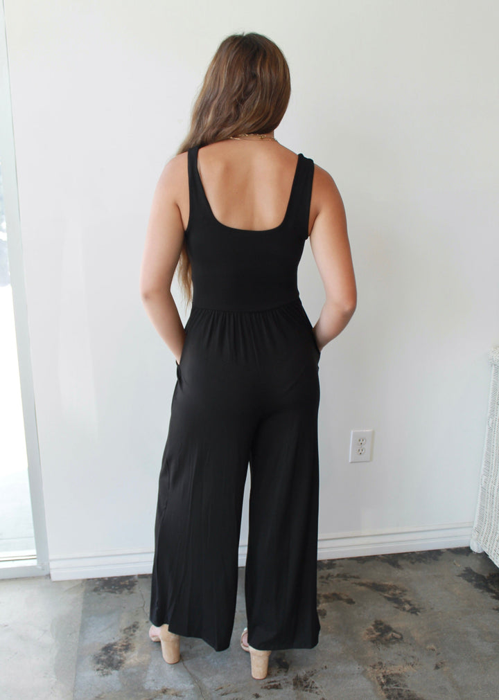 Woman wearing a black jumpsuit standing in a room with a white wall.
