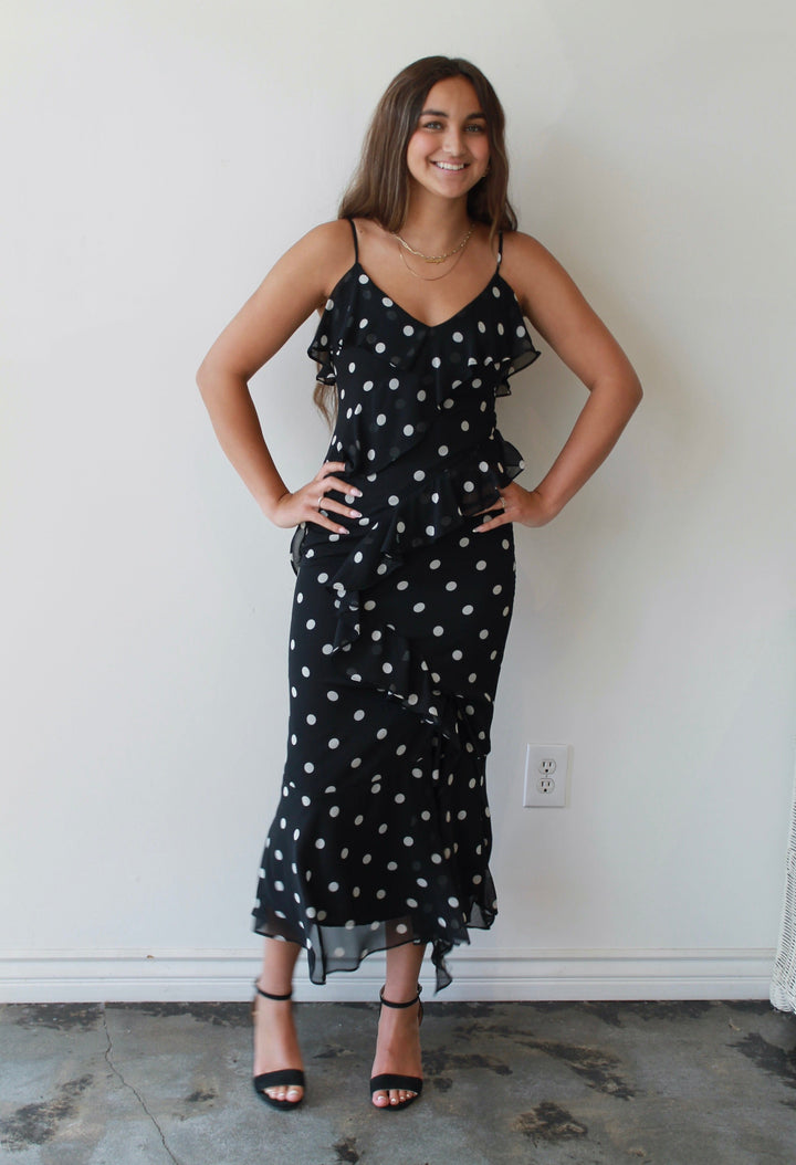 Woman wearing a black polka dot dress standing against a white wall.