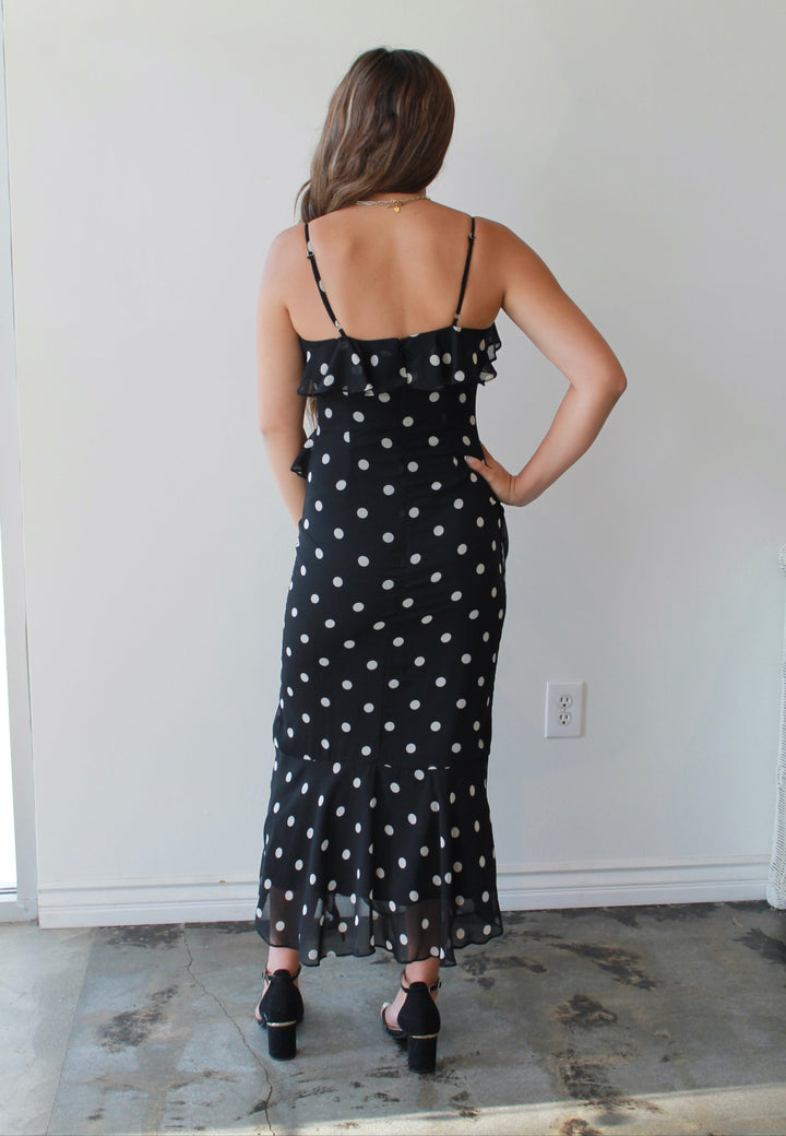 Woman wearing a black polka dot dress standing against a white wall.